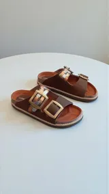 Sandalias de cuero color marrón, con diseño de dos tiras anchas con hebillas metálicas grandes y detalles pintados a mano. Suela de goma eva.