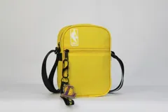 Morral pequeño de color amarillo con correa negra ajustable. Presenta el logo de la NBA en la parte superior y un relieve sutil de una pelota de baloncesto en el frente. Incluye un dije colgante con el logo de Los Angeles Lakers (letra 'L' con una pelota de baloncesto).