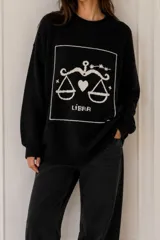 Sweater de punto negro con diseño oversize, cuello redondo y manga larga. Presenta un bordado artesanal en color crudo con el símbolo del signo zodiacal Libra, un corazón y estrellas dentro de un recuadro.