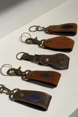 Llavero de cuero con gancho metálico tipo mosquetón y argolla, disponible en diversos diseños que incluyen bordados geométricos, costuras decorativas en contraste y apliques metálicos.