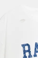 Camiseta de corte oversize confeccionada en algodón flamé, con cuello redondo y manga corta. Presenta un diseño con efecto desgastado, detalles de roturas en el tejido y estampado de texto a contraste en la parte frontal.
