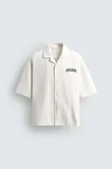 Camisa relaxed fit color blanco crudo, confeccionada en tejido de algodón con estructura. Cuello bowling y manga corta. Estampado frontal con la frase "ONLY DUST IS FOREVER" en negro. Cierre frontal de botonadura.