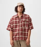 Camisa de franela de algodón con diseño a cuadros en tonos rojo y beige, de corte holgado relaxed, mangas cortas, cuello tradicional y cierre frontal con cremallera. Cuenta con un bolsillo aplicado en el pecho con detalle de botón metálico.