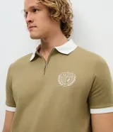Polo de manga corta color verde oliva, con cuello polo y puños en color blanco. Presenta cierre frontal parcial con cremallera y un escudo bordado en el pecho.