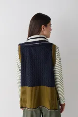 Chaleco de lana con cierre frontal completo, cuello alto y diseño bicolor en tonos marrón y celeste. Presenta una combinación de tejido trenzado y acanalado, con un corte asimétrico más largo en la parte posterior y aberturas laterales.