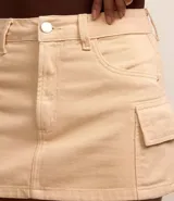 Pollera corta de sarga color beige claro, con cintura con trabillas y cierre de botón y cremallera. Presenta bolsillos cargo funcionales a los lados.