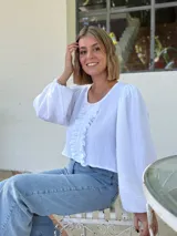 Blusa blanca de algodón con textura arrugada, cuello redondo, mangas largas abullonadas con puños elásticos y cierre frontal con botones y volantes.