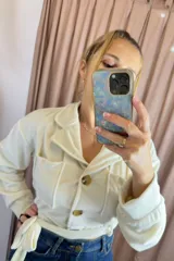 Campera tipo levita color crudo con textura efecto piel de durazno, cuello solapa, bolsillos frontales tipo cargo y cierre frontal con botones. Incluye un lazo ajustable en la cintura.