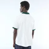 Camiseta tipo polo de manga corta, color blanco con cuello y ribetes en las mangas de color verde oscuro. Presenta un diseño gráfico estampado en el pecho con inscripciones y detalles en tono verde.