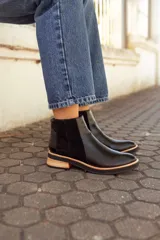 Botas tipo Chelsea de cuero negro, con paneles laterales elásticos, detalle de textura en el talón y suela plana con detalle en madera clara.