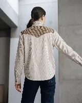 Camisa de manga larga con diseño de rayas verticales, confeccionada en algodón. Presenta un canesú superior en lana con estampado de cuadros en tonos tierra, cuello alto con volados y cierre frontal con botones.