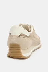 Zapatilla deportiva tipo running en tonos beige y crema, con combinación de materiales como gamuza y tela. Presenta pespuntes decorativos en el cuerpo, cierre de cordones y punta redonda. La suela tiene una altura de 3 cm.