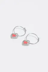 Aros de metal tipo argolla con dije colgante en forma de corazón esmaltado en color blanco y rosado, con detalles de letras estampadas.