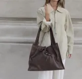Bolso tipo shopper de cuero color marrón chocolate, con diseño amplio y cierre de cordón ajustable en la parte frontal. Cuenta con doble asa de hombro, forro interior, bolsillo interno y externo, y llavero incorporado.
