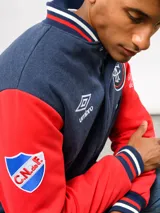 Campera bomber azul marino estilo retro, con cierre frontal de botones a presión, cuello, puños y ruedo con tejido acanalado tricolor. Presenta el logo de Umbro y el escudo del Club Nacional de Football bordados en el pecho, junto con el año 1899 y parches decorativos en las mangas.