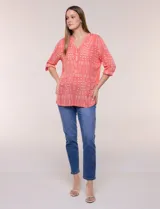 Blusa turquesa de silueta suelta confeccionada en tejido liviano, con escote en V, pequeña cartera frontal con botones decorativos y mangas 3/4 con presilla para arremangar. Presenta patrón estampado geométrico tipo tie-dye y ruedo curvo.