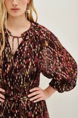 Vestido midi de corte holgado con estampado ikat en tonos bordó y tierra. Presenta cuello redondo con lazos, mangas largas abullonadas con puños elásticos y falda con volados escalonados. Incluye un cinturón ancho de estilo rústico con tachas y flecos.