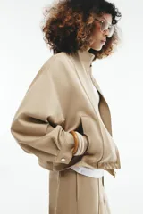 Cazadora bomber de color beige con cuello subido, manga larga y bolsillos delanteros. Cuenta con cierre frontal de cremallera metálica y cordón ajustable en el dobladillo.