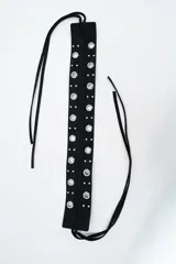 Cinturón ancho de color negro con diseño de ojales metálicos y tachas decorativas. Cuenta con tiras largas para anudar.