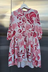 Vestido de tela ligera y fluida con estampado floral en tonos rosados y rojos sobre fondo blanco. Presenta cuello redondo tipo mao, cierre frontal con botones, mangas tres cuartos y corte con volados que generan una silueta relajada y con movimiento.