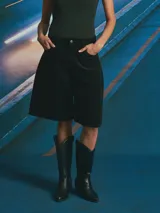 Bermuda de corte amplio y tiro alto, confeccionada en tejido de pana color negro. Presenta diseño de cinco bolsillos, cierre frontal con botón y bragueta, y largo por encima de la rodilla.