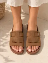 Sandalias de gamuza color marrón, con diseño de una sola tira ancha decorada con flecos y una hebilla metálica lateral ajustable. Cuenta con plantilla anatómica.