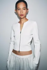 Chaqueta de punto liso color blanco, con cuello redondo, manga larga acabada con cremalleras en los puños y cierre frontal con cremallera.