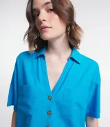 Camisa de manga corta confeccionada en mezcla de viscosa y lino color turquesa. Presenta cuello camisero con escote en V, bolsillos frontales de parche y cierre frontal con botones geométricos de efecto carey.