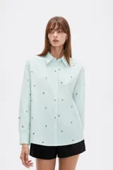 Blusa de corte holgado con cuello camisero y cierre frontal de botones. Presenta un diseño de rayas verticales finas en color azul y blanco, decorado con pequeños bordados dispersos.