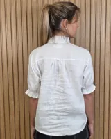 Camisa de denim de algodón celeste, con cierre frontal de botones, cuello alto con volados y mangas cortas abullonadas con puños elastizados fruncidos.