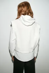 Camisa blanca de manga larga con un top de sudadera gris claro superpuesto, que incluye capucha ajustable con cordones. El bajo de la camisa tiene aberturas laterales.