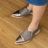 Zapato tipo mocasín de cuero vacuno en tono gris topo, con puntera reforzada en cuero metalizado color bronce. Presenta un diseño de corte lateral y suela plana con pequeña elevación en el talón.