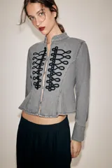 Chaqueta de denim gris con cuello subido y manga larga. Presenta un diseño entallado con bajo en evasé y un llamativo bordado frontal de alamares en color negro. Cierre frontal con corchetes ocultos.