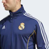Campera deportiva Adidas de hombre, color azul marino, inspirada en la equipación del Real Madrid temporada 99/00. Presenta cuello alto, cierre frontal, tres franjas blancas en las mangas y ribetes amarillos en los costados. Lleva el escudo del Real Madrid y el logo de Adidas bordados en el pecho.