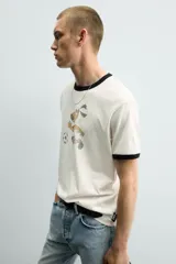 Camiseta de algodón color crudo con cuello y bordes de mangas en contraste negro. Presenta un estampado frontal de estilo vintage con el personaje Striker, mascota de la Copa Mundial de la FIFA 1994, jugando al fútbol.