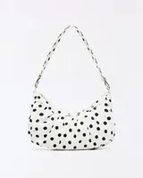 Cartera tipo hobo de diseño arqueado con estampado de lunares negros sobre fondo blanco. Cuenta con cierre superior de cremallera, tiras fruncidas decorativas en la base y una correa de hombro ajustable con argollas metálicas laterales.