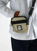 Bolso tipo morral pequeño de la marca Obey, confeccionado en lona de poliéster. Presenta un diseño bicolor en tonos beige y negro, con un parche frontal del logo de la marca en PVC. Cuenta con cierre metálico, bolsillo interno, correa regulable y herrajes plásticos.