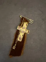 Pin decorativo con forma de cruz dorada de estilo vintage, adornada con piedras incrustadas. La pieza incluye una base de tela de terciopelo desflecada, una cadena metálica y un pequeño dije colgante.