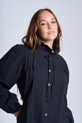 Camisa de color azul con cuello alto decorado con volados y lazo de ajuste. Presenta tablas decorativas en los hombros, cierre frontal con botones de nácar y mangas largas con puños elásticos que generan un efecto abullonado.