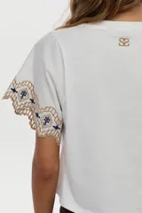 Blusa blanca de jersey con mangas cortas amplias de tela plana, decoradas con bordados geométricos en tonos marrón y azul marino.