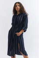 Vestido camisero midi de lino color azul marino con rayas verticales blancas, corte holgado, cuello camisero, mangas 3/4 con puños elásticos y lazo en la cintura.