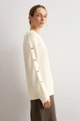 Sweater de punto color blanco natural, con cuello redondo y mangas largas adornadas con botones dorados a lo largo de las costuras exteriores.