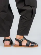 Sandalias planas de cuero negro estilo pescador, con tiras anchas entrelazadas en el empeine, suela marrón y tira ajustable con hebilla metálica en el tobillo.