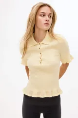 Top entallado en punto suave acanalado de color amarillo pastel, con cuello camisero, manga corta y tapeta con botones metálicos. Presenta detalles de volados en los puños y el bajo.