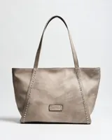 Cartera tipo tote de símil cuero color taupe, con doble asa de hombro, detalles de tachas plateadas en los laterales frontales y placa metálica con logo grabado de la marca XL Extra Large en el centro.