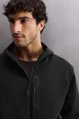 Sudadera con capucha y cierre de cremallera frontal, color negro, confeccionada en tejido técnico compacto de algodón elástico. Presenta cuello subido, bolsillos frontales con cremallera y acabado termofijado, y puños y bajo en rib del mismo tejido.