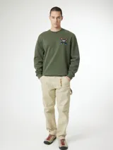 Sudadera de cuello redondo color verde oliva, con detalle de bordado en forma de X con patrón geométrico multicolor en el pecho.