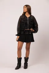 Campera bomber negra con transparencias, confeccionada en gasa con seda labrada. Presenta cierre frontal metálico, bolsillos cargo frontales con tachas y tiras colgantes, mangas con volumen y puños y cuello de rib elástico.