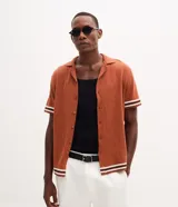 Camisa de punto jacquard color naranja, con cuello solapa, manga corta y cierre frontal con botones. Presenta detalles de rayas horizontales en contraste en el ruedo y en los puños de las mangas.