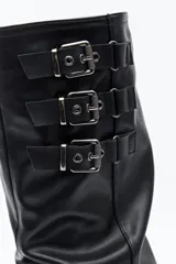 Bota de caña alta color negro, con diseño utilitario, puntera redondeada y taco bajo. Presenta un efecto fruncido en la caña y tres hebillas metálicas decorativas.